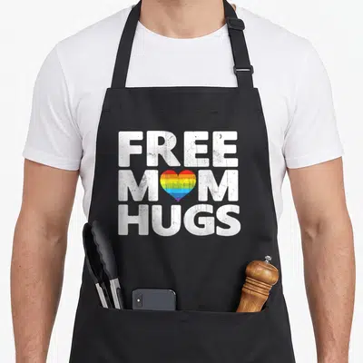 Free gay pride apron - Distressed white text FREE MOM HUGS with rainbow heart replacing O in mom — shown on a black cotton canvas apron against a white background