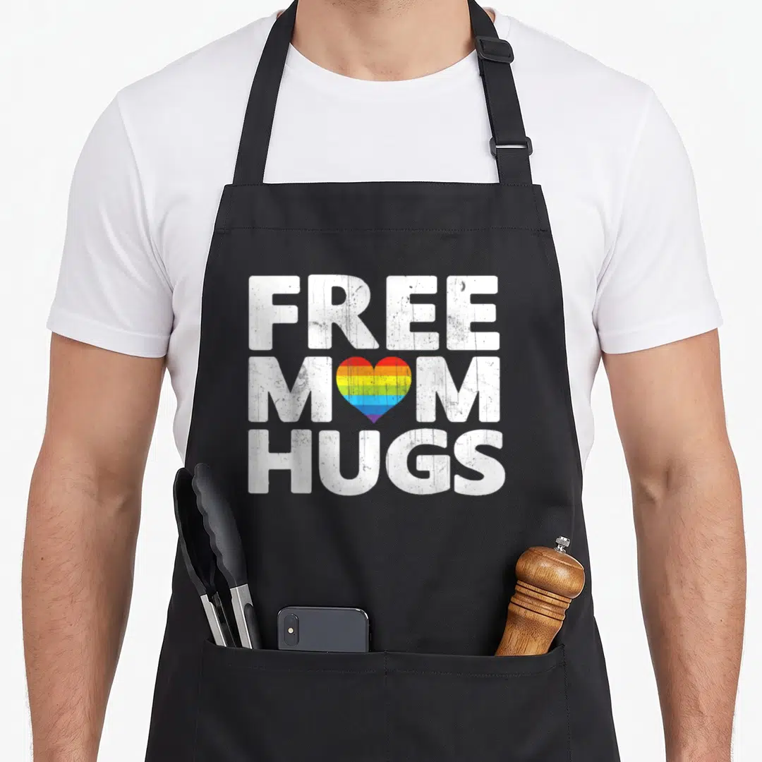 Free gay pride apron - Distressed white text FREE MOM HUGS with rainbow heart replacing O in mom — shown on a black cotton canvas apron against a white background