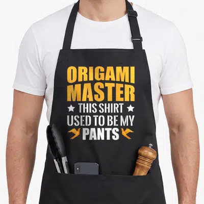 Origami master apron - Orange origami birds flanking white text reading This Shirt Used To Be My Pants below stacked — shown on a black cotton canvas apron against a white background