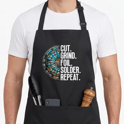 Grind stained apron - Semi-circle stained glass in blue orange white next to stacked text Cut Grind Foil Solder Repeat — shown on a black cotton canvas apron against a white background