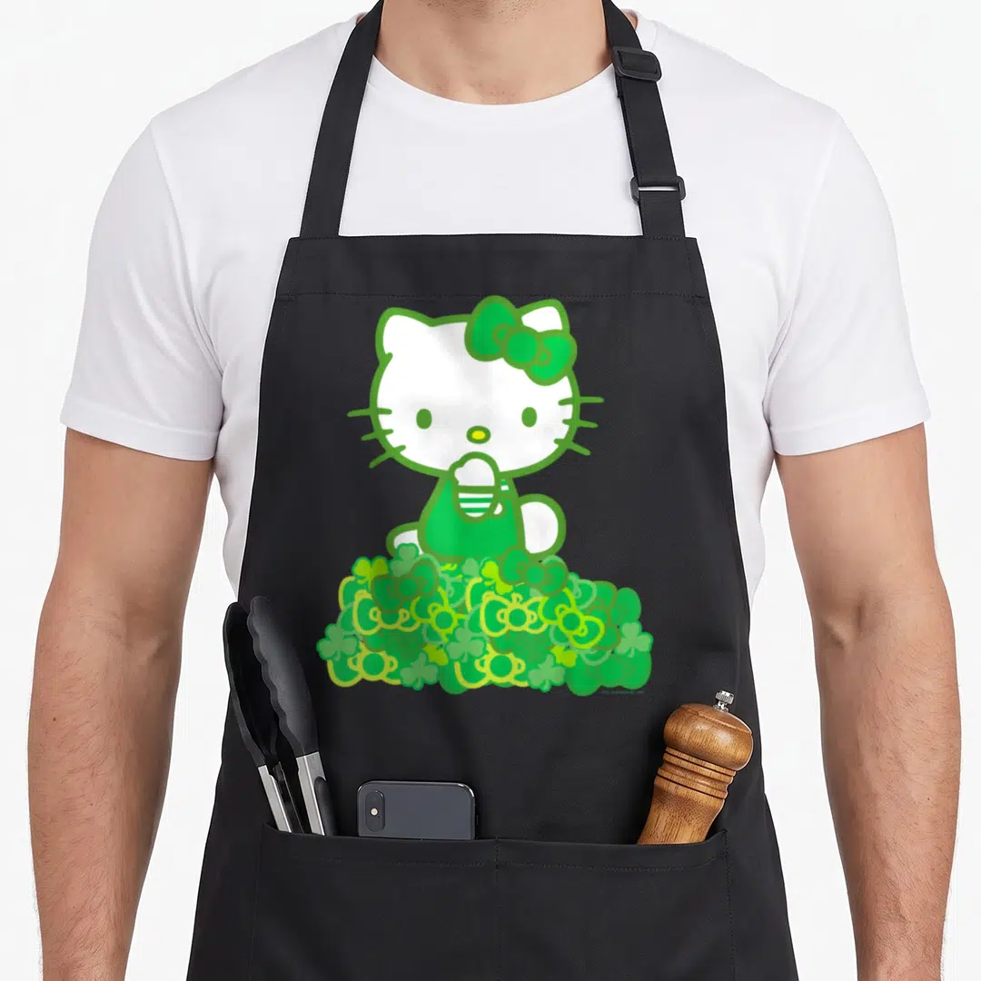 Funny clover bow apron - Hello Kitty in green and white sitting on a pile of shamrocks and bows — shown on a black cotton canvas apron against a white background