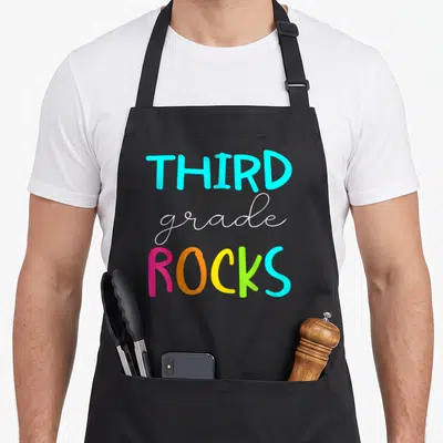 Third cute teacher aprons - Stacked cyan white pink orange yellow lime green text reading THIRD grade ROCKS — shown on a black cotton canvas apron against a white background