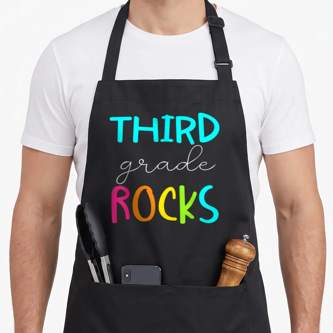 Third cute teacher aprons - Stacked cyan white pink orange yellow lime green text reading THIRD grade ROCKS — shown on a black cotton canvas apron against a white background