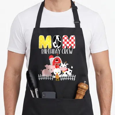 Barnyard crew aprons - MOM BIRTHDAY CREW text above barn and three animals behind white fence — shown on a black cotton canvas apron against a white background