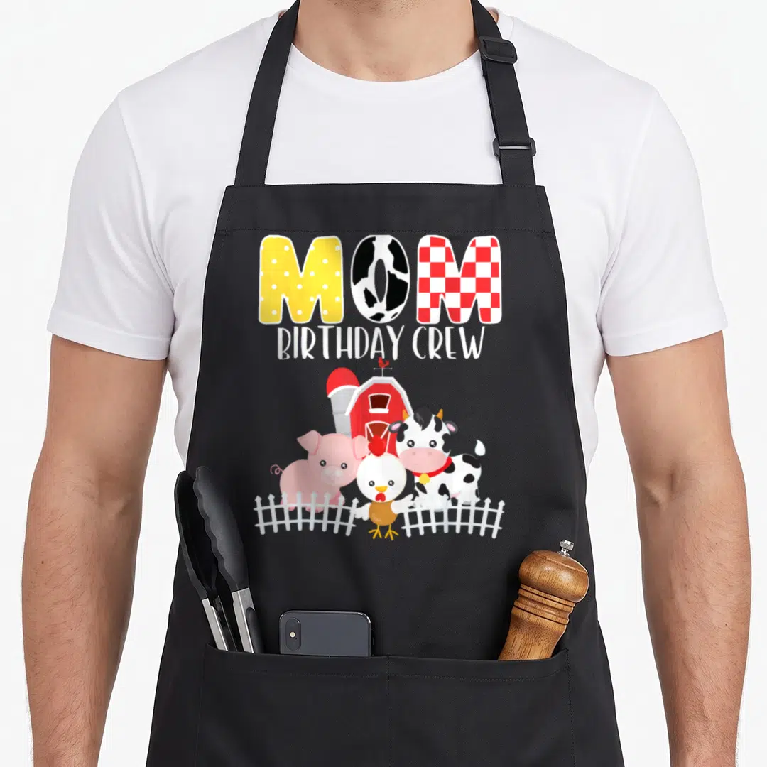 Barnyard crew aprons - MOM BIRTHDAY CREW text above barn and three animals behind white fence — shown on a black cotton canvas apron against a white background