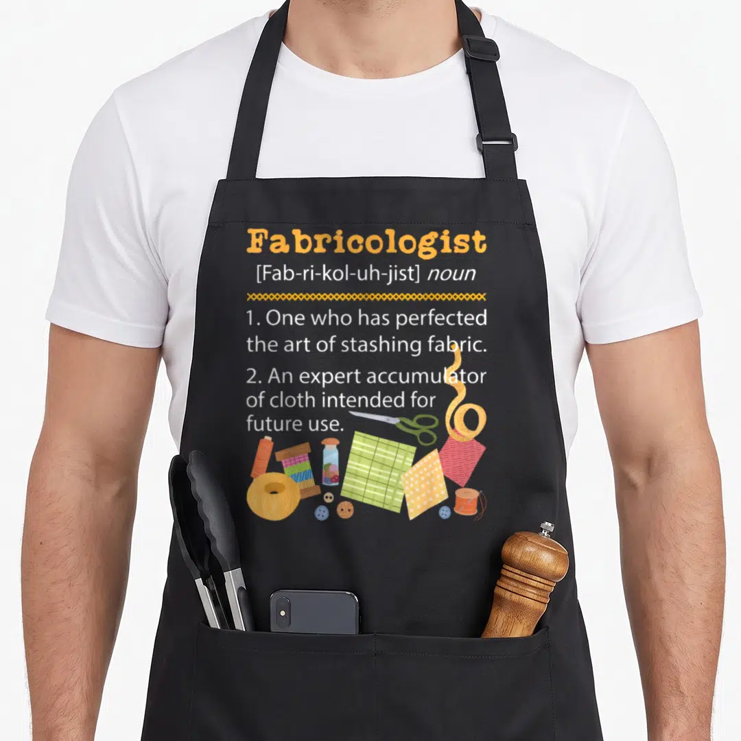 Sewer needlework apron - Fabricologist dictionary definition with cartoon scissors, thread, and fabric scraps — shown on a black cotton canvas apron against a white background