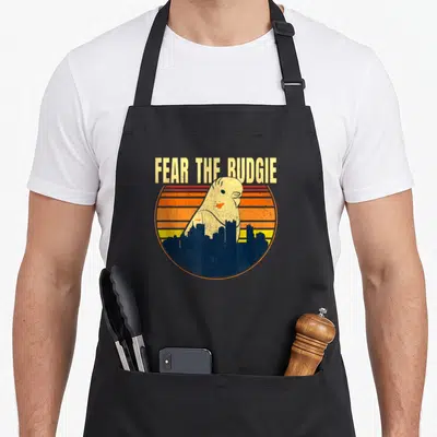 Parrot crazy parakeet man apron - FEAR THE BUDGIE text above budgie silhouette and dark blue city skyline inside — shown on a black cotton canvas apron against a white background