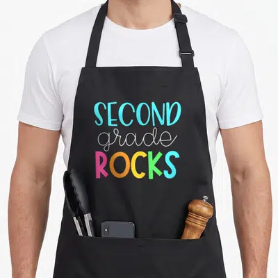 Teacher 2nd apron - Stacked SECOND grade ROCKS text in blue white pink orange yellow and green typography — shown on a black cotton canvas apron against a white background