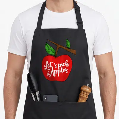 Pick apple apron - White Let's pick Apples script on a red apple with brown branch and two green leaves — shown on a black cotton canvas apron against a white background
