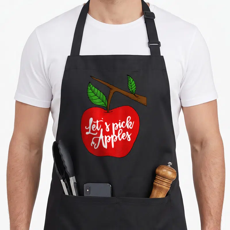 Pick apple apron - White Let's pick Apples script on a red apple with brown branch and two green leaves — shown on a black cotton canvas apron against a white background