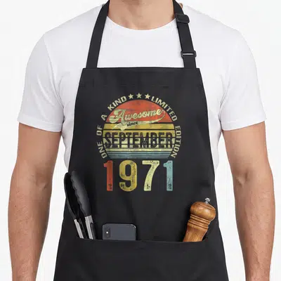 September 50th birthday apron - Retro sun graphic with Awesome since SEPTEMBER 1971 text in red yellow blue and beige — shown on a black cotton canvas apron against a white background