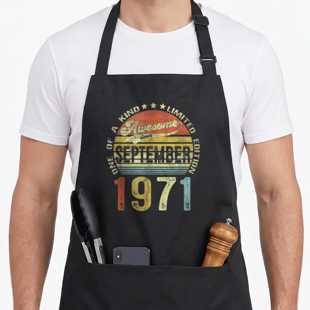 September 50th birthday apron - Retro sun graphic with Awesome since SEPTEMBER 1971 text in red yellow blue and beige — shown on a black cotton canvas apron against a white background