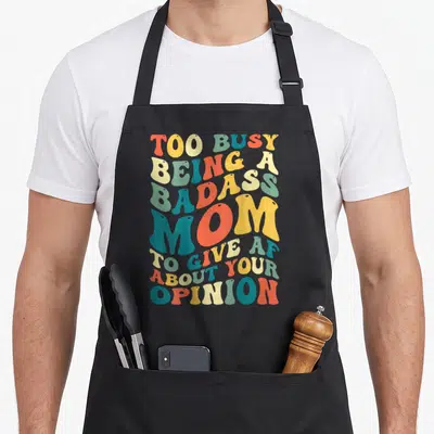 Badass busy apron - Retro bubbly text reading TOO BUSY BEING A BADASS MOM TO GIVE AF ABOUT YOUR OPINION in orange — shown on a black cotton canvas apron against a white background