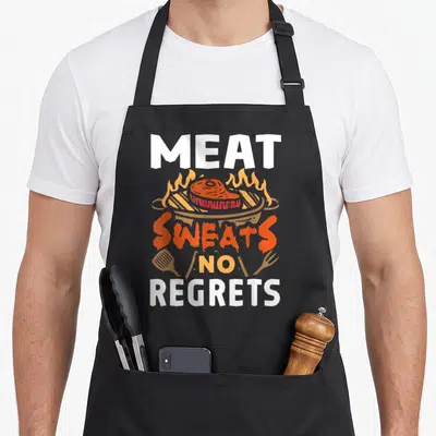 Sweat barbecue apron - Cartoon steak on grill with Meat Sweats No Regrets text in white and orange — shown on a black cotton canvas apron against a white background