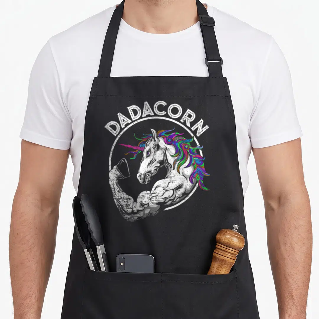 Unicorn muscle apron - Muscular unicorn flexing inside white circle with Dadacorn text arching overhead — shown on a black cotton canvas apron against a white background