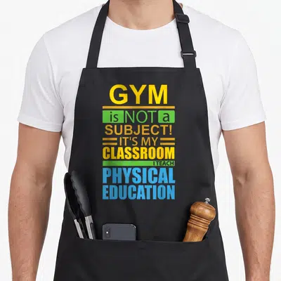 Gym education apron - Typography reading GYM is NOT a SUBJECT IT'S MY CLASSROOM I TEACH PHYSICAL EDUCATION with green — shown on a black cotton canvas apron against a white background