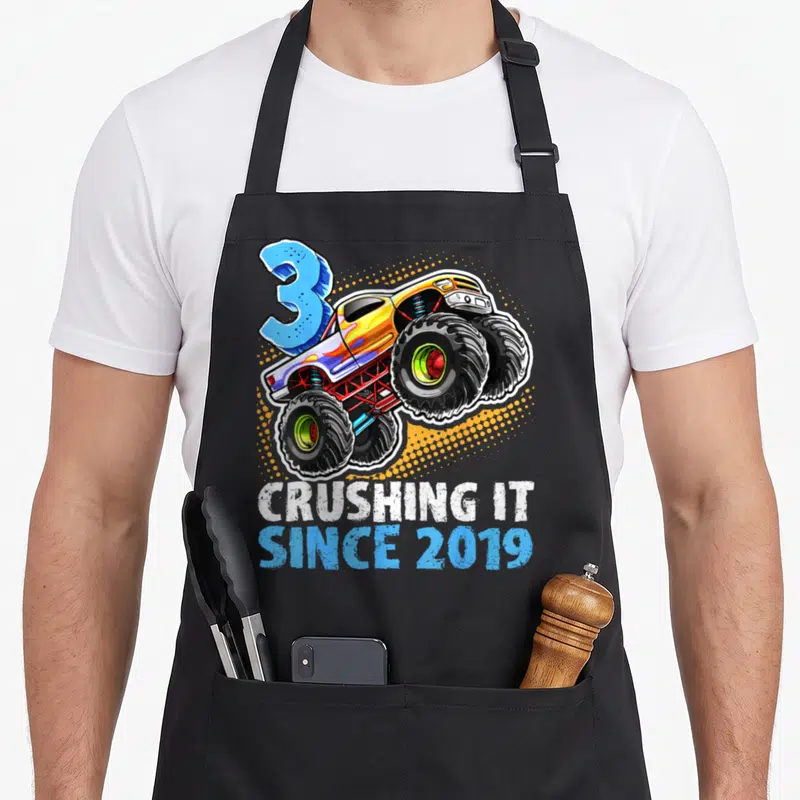 Crushing monster truck apron - Blue number 3 monster truck over orange halftone dots with white and blue text — shown on a black cotton canvas apron against a white background