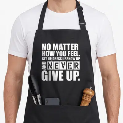 Motivation ultimate apron - White stacked typography reading NO MATTER HOW YOU FEEL GET UP DRESS UP SHOW UP AND NEVER — shown on a black cotton canvas apron against a white background