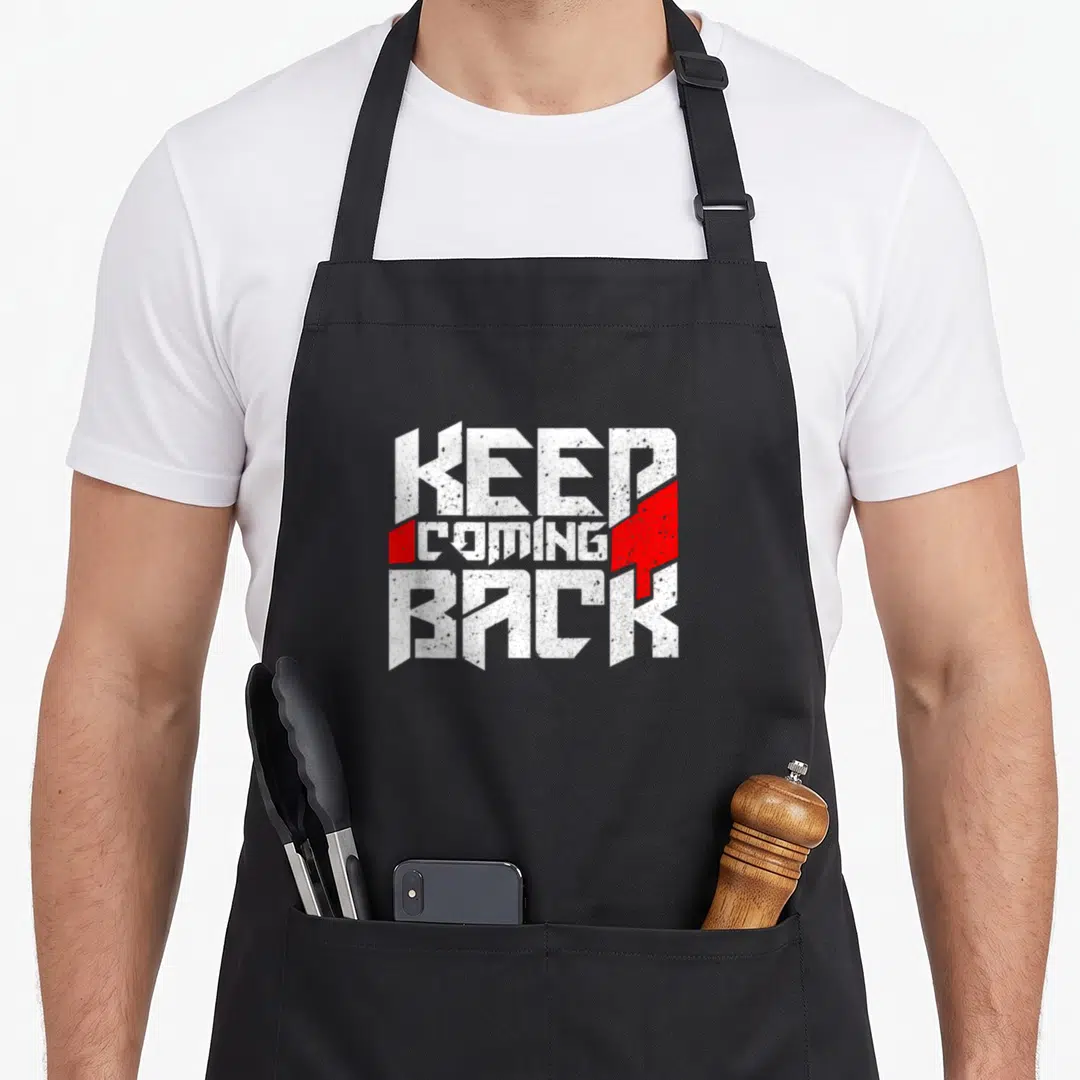 Coming celebrate apron - White distressed text KEEP COMING BACK flanked by red geometric shapes — shown on a black cotton canvas apron against a white background