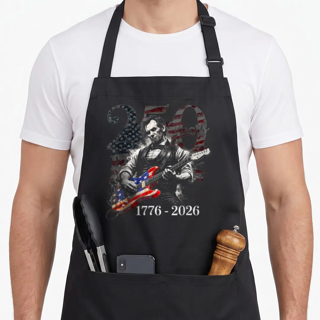 250th day july apron - Abraham Lincoln playing flag-patterned guitar with 250 Years text and 1776-2026 dates on grey — shown on a black cotton canvas apron against a white background