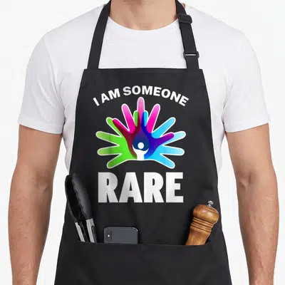 Funny rare disease awareness apron - I Am Someone Rare text above pink blue green purple hands with central white figure — shown on a black cotton canvas apron against a white background