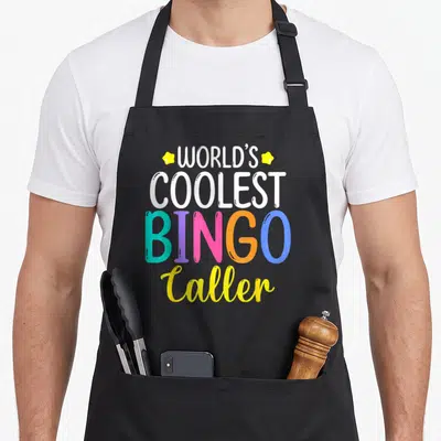 Player coolest bingo aprons - Stacked text reading World's Coolest Bingo Caller with multi-colored letters and yellow — shown on a black cotton canvas apron against a white background