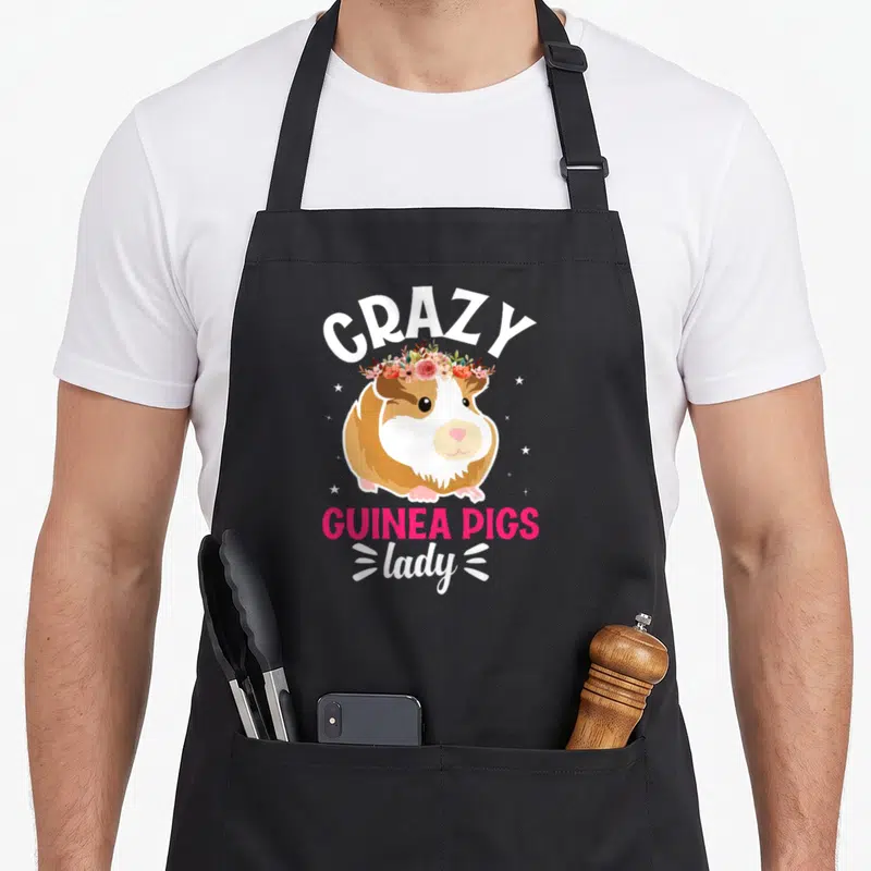 Pig crazy guinea lady apron for hosting - Cartoon guinea pig with pink flower crown and white and pink text surrounded — shown on a black cotton canvas apron against a white background