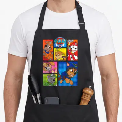 Pup box apron - Five comic panels of puppies with PAW PATROL shield logo on black background — shown on a black cotton canvas apron against a white background