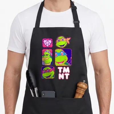 Portrait box apron - Four rounded squares with turtle heads and heart, plus TM NT text in green pink blue white — shown on a black cotton canvas apron against a white background
