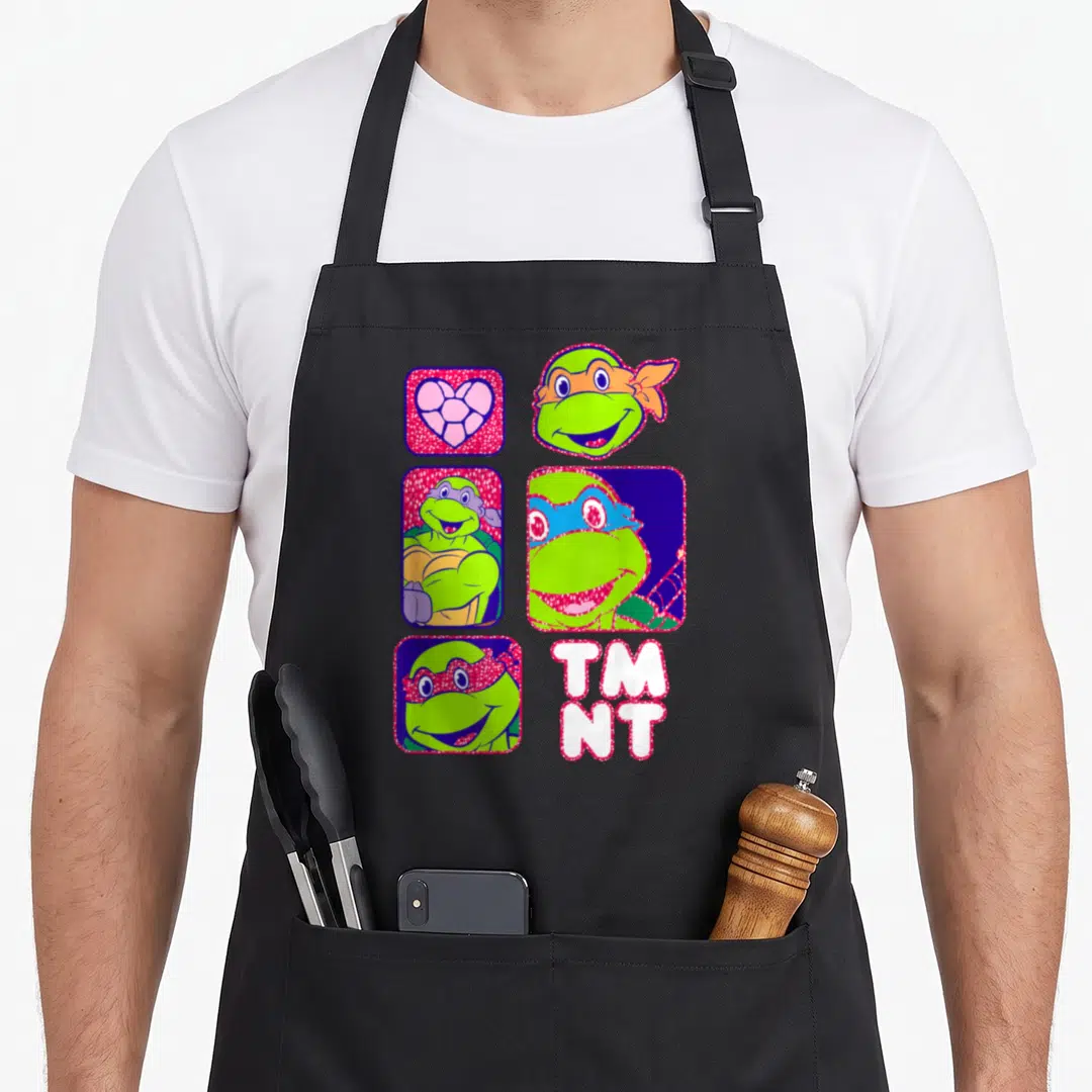 Portrait box apron - Four rounded squares with turtle heads and heart, plus TM NT text in green pink blue white — shown on a black cotton canvas apron against a white background