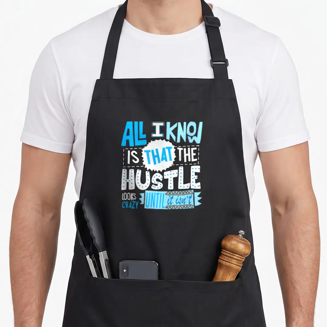 Hustle blue apron - Stacked blue and white text reading All I Know Is That The Hustle Looks Crazy Until It Isn't — shown on a black cotton canvas apron against a white background