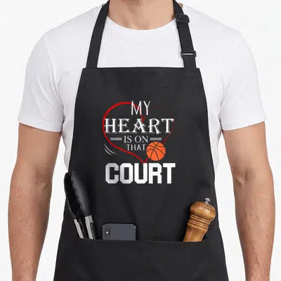 Court basketball dad apron - Red heart outline enclosing white text My Heart Is On That Court with orange basketball — shown on a black cotton canvas apron against a white background