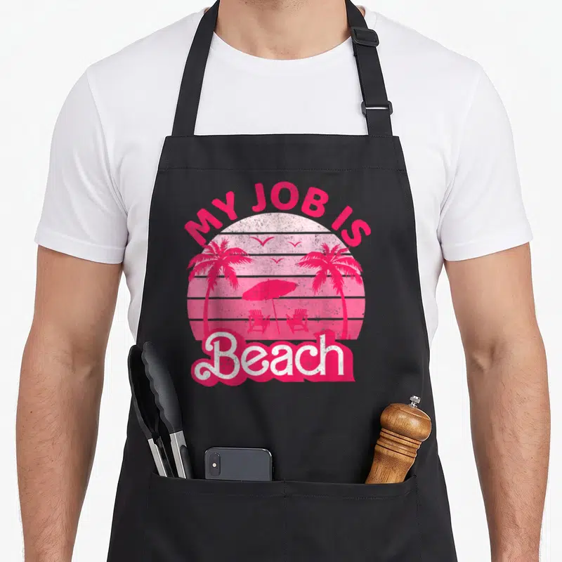 Vintage beach apron - Pink retro sunset with palm trees, umbrella, chairs, and MY JOB IS Beach text — shown on a black cotton canvas apron against a white background