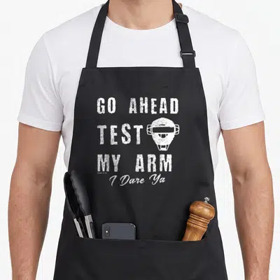 Fastpitch catcher apron - White distressed catcher mask with stacked text GO AHEAD TEST MY ARM — shown on a black cotton canvas apron against a white background