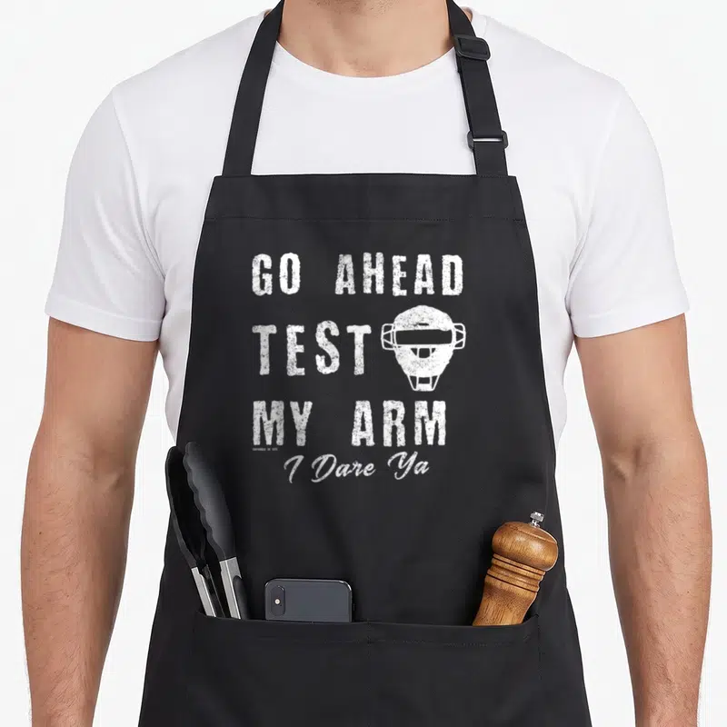 Fastpitch catcher apron - White distressed catcher mask with stacked text GO AHEAD TEST MY ARM — shown on a black cotton canvas apron against a white background