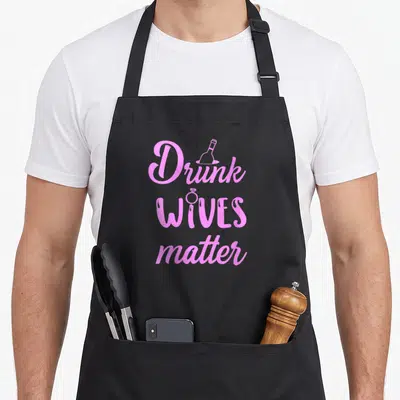 Drunk champagne apron - Pink stacked text Drunk WIVES matter with wine bottle and ring graphics on black background — shown on a black cotton canvas apron against a white background