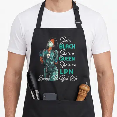 Lpn queen apron - Realistic nurse in teal scrubs with stacked text She's Black She's a Queen She's an LPN — shown on a black cotton canvas apron against a white background