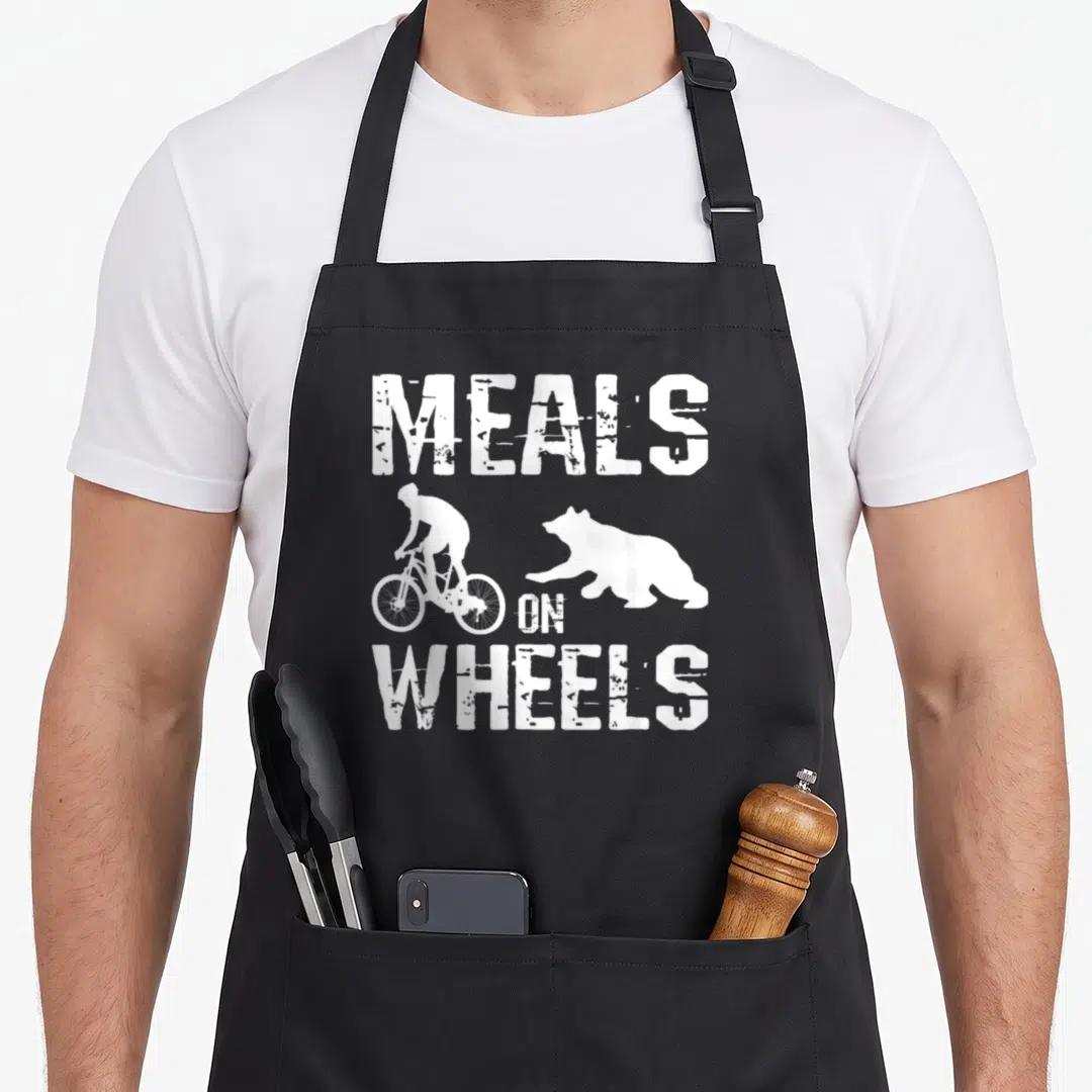 Meal wheel apron - White distressed text MEALS ON WHEELS with cyclist and bear silhouettes on black fabric — shown on a black cotton canvas apron against a white background