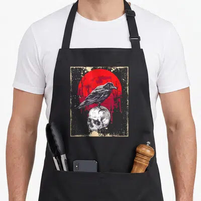 Skull moon gothic apron - Raven perched on skull with barbed wire and red sun in distressed beige frame — shown on a black cotton canvas apron against a white background