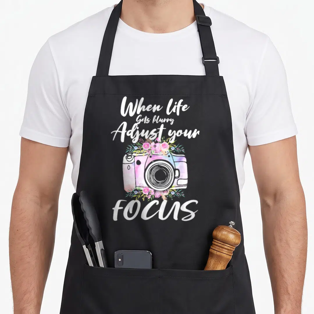 Vintage camera photographer apron - Watercolor camera with pink flowers and text When Life Gets blurry Adjust your FOCUS — shown on a black cotton canvas apron against a white background