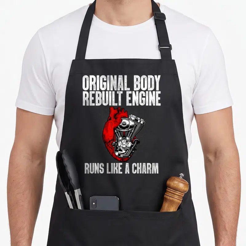 Surgery open apron - Red heart merged with silver engine and white text Original Body Rebuilt Engine Runs Like A Charm — shown on a black cotton canvas apron against a white background