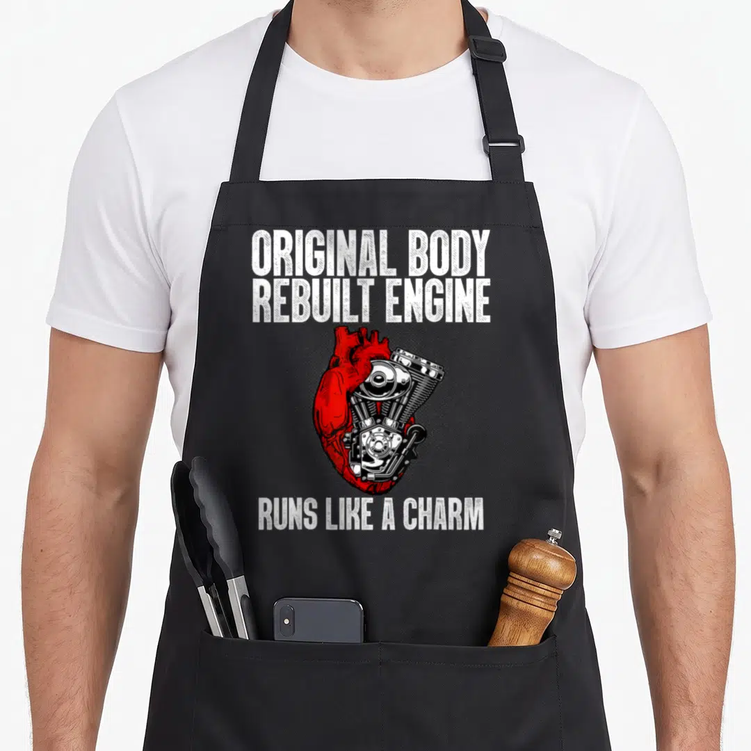 Surgery open apron - Red heart merged with silver engine and white text Original Body Rebuilt Engine Runs Like A Charm — shown on a black cotton canvas apron against a white background
