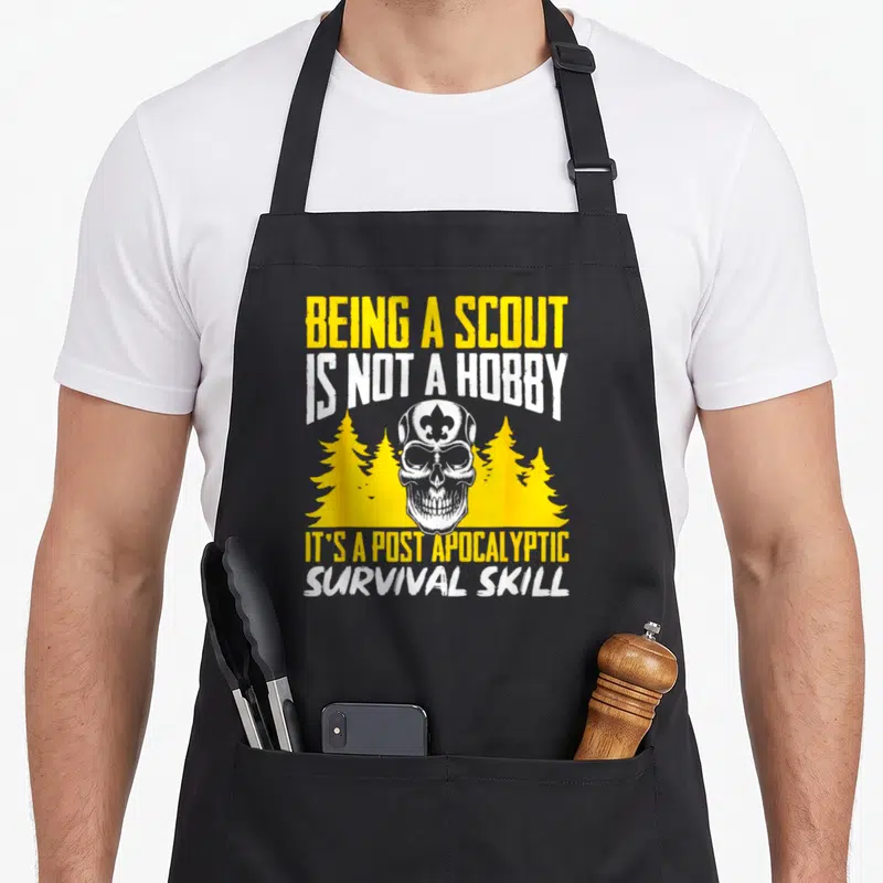 Scout outdoor camping apron - Distressed skull with fleur-de-lis and yellow pine trees under survival skill quote — shown on a black cotton canvas apron against a white background