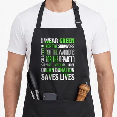 Modern organ apron - Stacked I WEAR GREEN text over a green awareness ribbon representing organ donation support — shown on a black cotton canvas apron against a white background
