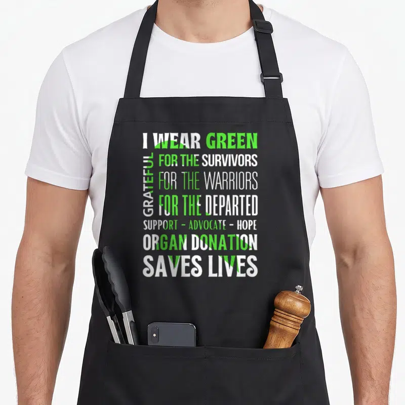 Modern organ apron - Stacked I WEAR GREEN text over a green awareness ribbon representing organ donation support — shown on a black cotton canvas apron against a white background