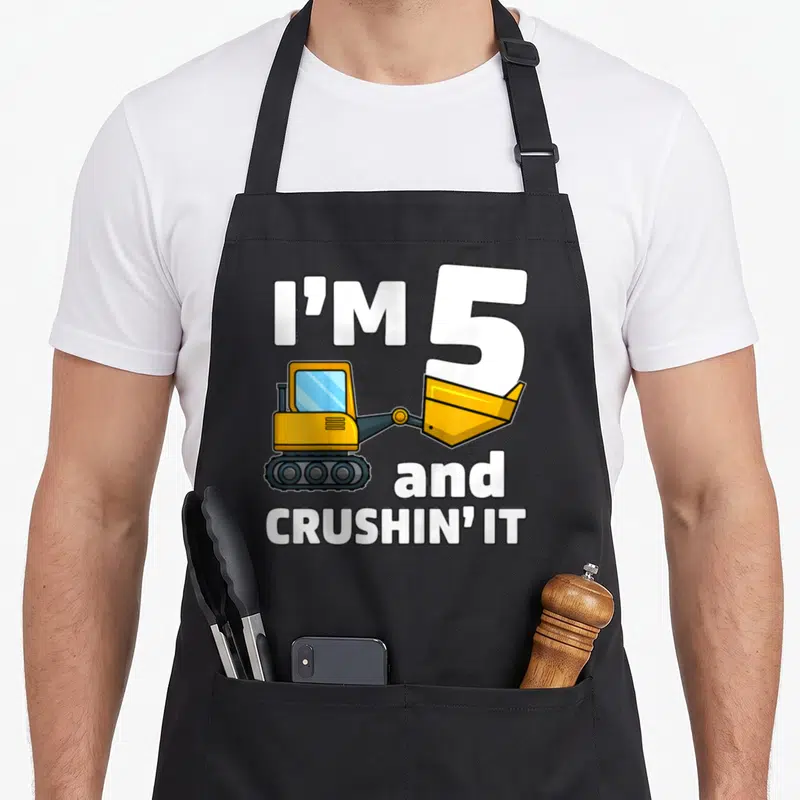 Construction 5th truck apron - Cartoon excavator lifting number 5 with text I'm 5 and crushing it — shown on a black cotton canvas apron against a white background
