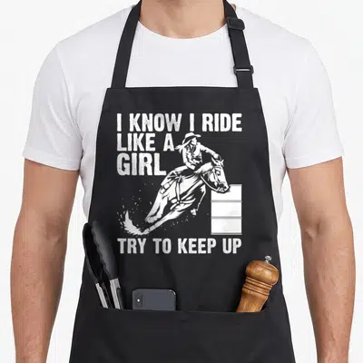 Ride racing apron - White distressed text I know I ride like a girl try to keep up above a horse jumping a barrel — shown on a black cotton canvas apron against a white background