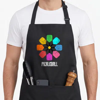 Pickleball rainbow apron - Eight colorful paddles radiating from yellow ball with PICHLEBALL text below — shown on a black cotton canvas apron against a white background