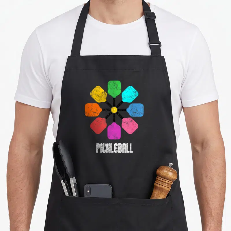 Pickleball rainbow apron - Eight colorful paddles radiating from yellow ball with PICHLEBALL text below — shown on a black cotton canvas apron against a white background