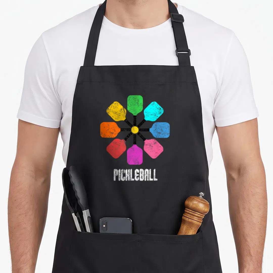 Pickleball rainbow apron - Eight colorful paddles radiating from yellow ball with PICHLEBALL text below — shown on a black cotton canvas apron against a white background